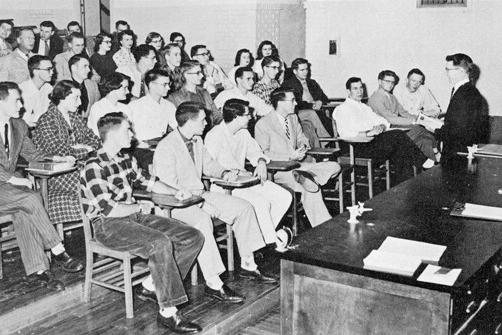 这是一张黑白照片，左边是一位教授站在讲堂前，学生们坐在台阶上错开的桌子旁.